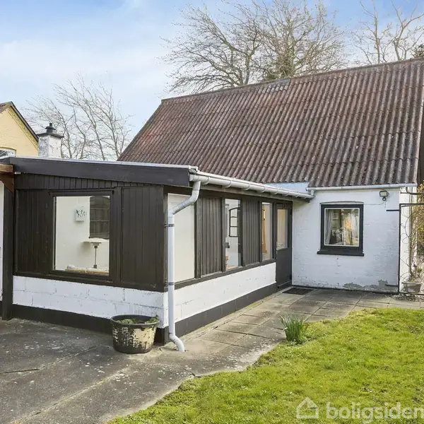 Hus med hvid facade og mørkt træværk ses fra gårdsplads. Taget er brunt. En græsplæne og betonsti omgiver huset. Baggrunden har nøgne træer under en blå himmel.