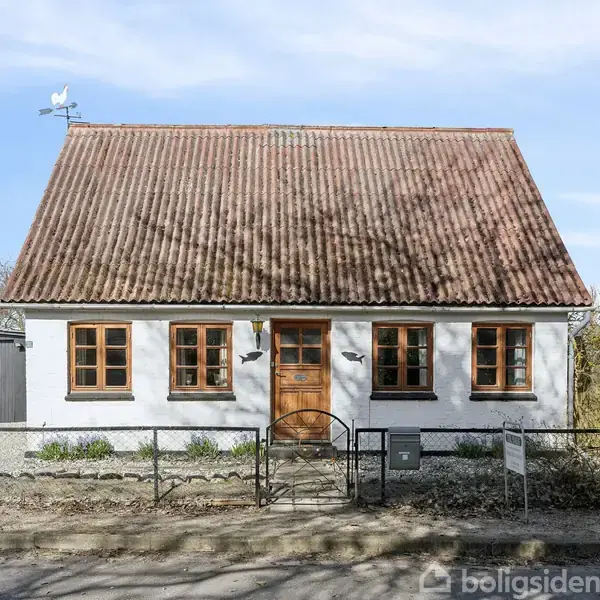 Et hvidt hus med rødt tegltag står foran en grusvej, omgivet af en lav hegn, træer og en mindre bygning til venstre.