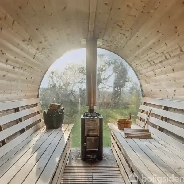 En træsaunaformet struktur indeholder bænke langs væggene. En brændeovn står i midten med udsigt til natur gennem et vindue i baggrunden. Ingen tekst til stede.