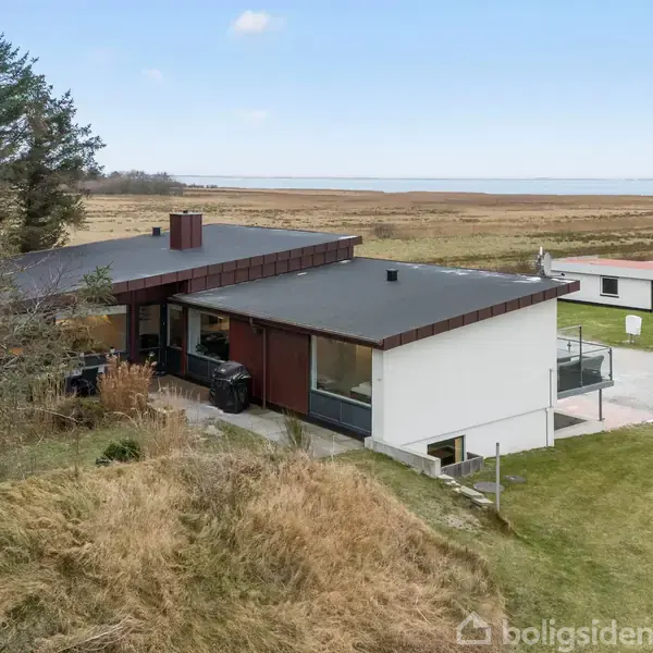 Et moderne hus med fladt tag og store vinduer står i en åben, naturlig omgivelser tæt på havet med lidt træbevoksning.