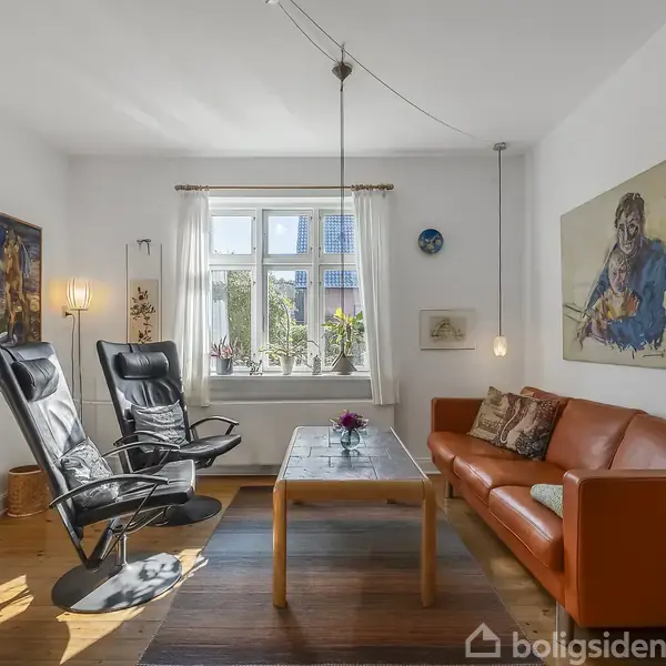 En stue med en orange sofa og to sorte lænestole omkring et træbord. Sollys strømmer gennem vinduet, og vægkunst pryder rummets hvide vægge. En bogreol står til højre.