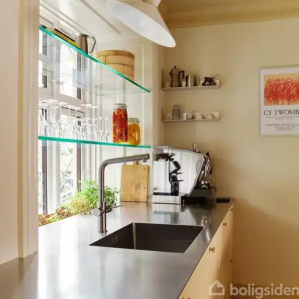 Køkkenvask med armatur står på en skinnende bordplade. Rundt om er der hylder med glas og konserveringsglas. En kaffemaskine ses ved siden af vasken. Plakat med "CY TWOMBLY" på væggen.