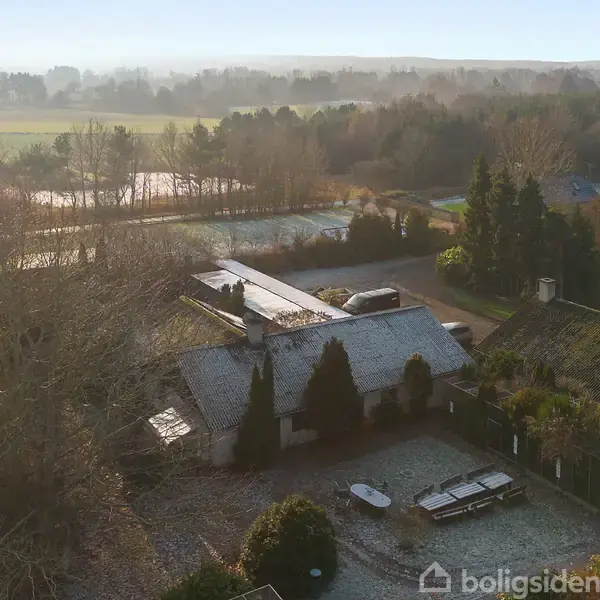 Huset er dækket af rimfrost, omkranset af træer og buske. Sne og frost pryder omgivelserne i det landlige område med marker og skove i baggrunden.