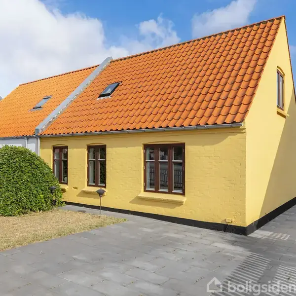 Gult murstenshus med orange tegltag står på flisebelagt gårdsplads. En lille græsrabat med en grøn busk er ved siden af huset. Klar blå himmel i baggrunden.