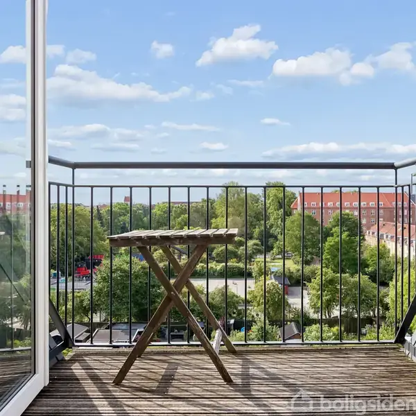 En lille balkon med et sammenklappeligt bord står åben ud mod en udsigt med frodige træer og røde tage under en blå himmel med spredte skyer.