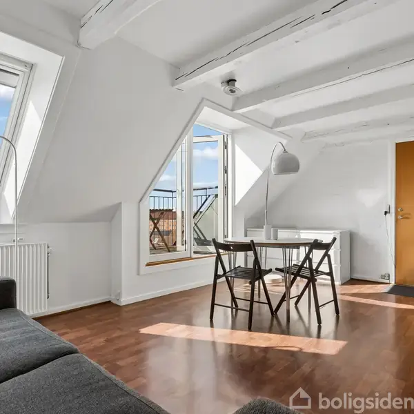 Et lyst, rummeligt værelse med trægulv indeholder en sort sofa og et lille spisebord med to stole. Skråvinduer og en glasdør fører ud til en altan med udsigt.