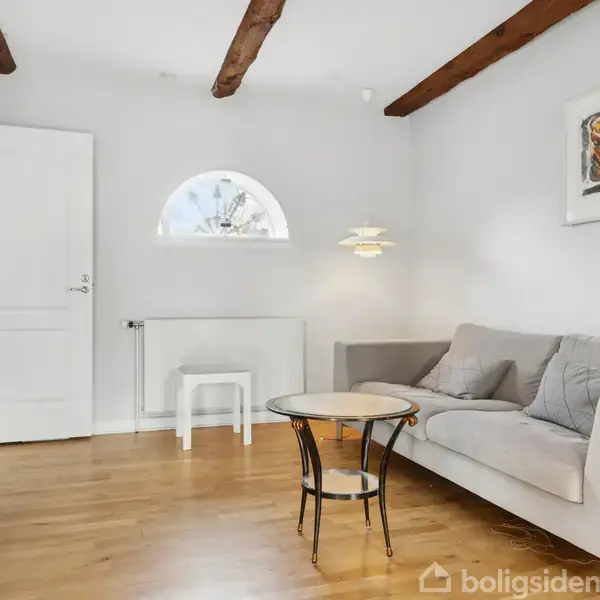 En stue indeholder en grå sofa ved siden af et rundt glasbord. Trægulvet suppleres af synlige loftsbjælker. En fladskærm står på et møbel til venstre, og der hænger kunst på væggen.