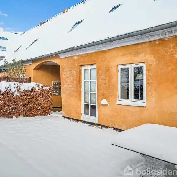 Et gult hus med hvide døre og vinduer står i et sneklædt område. Hækken er dækket af sne, og et garageanlæg ses i baggrunden.