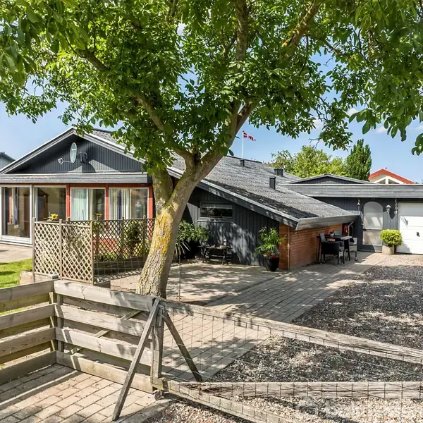 Et moderne parcelhus med sort facade og hvide vinduer står stille omkranset af en grusbelagt indkørsel og grønne buske, mens en stor træterrasse tilbyder udendørs opholdsplads.