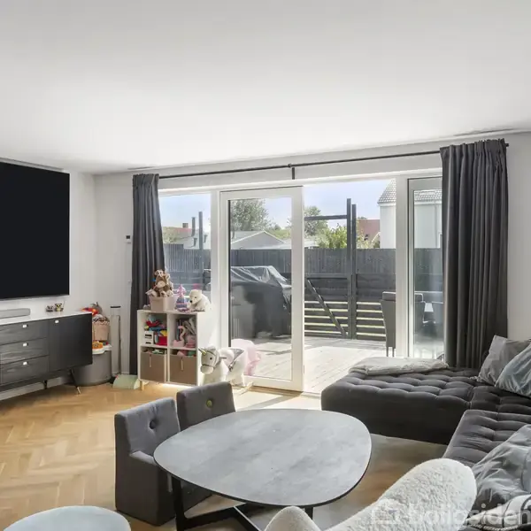 Stue med en stor, mørkegrå sofa, rundt sofabord og sort TV på væggen. Skydedøre fører ud til en terrasse med grill og haveudsigt. Parketgulve tilføjer varme.