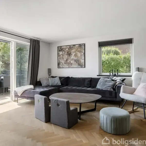 Stue med sort sofa og rundt sofabord foran vinduer mod terrasse; skænk med dekorationer under vægkunst. Lyse vægge og trægulv skaber en moderne atmosfære.