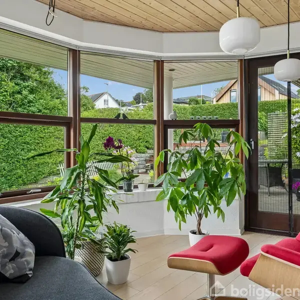 Stue møbleret med sort sofa og røde stole, omgivet af planter. Store vinduer giver udsigt til grøn have. Moderne lamper hænger fra loftet, og en glasdør fører ud til terrassen.