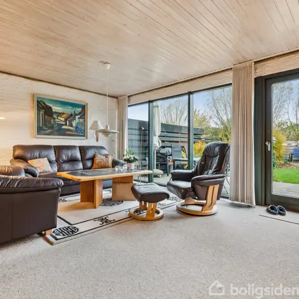 En stue med sorte lædermøbler placeret omkring et træbord, handling i et rum med maleri på væggen, store vinduer og glasdør, der fører til en have på udsiden.