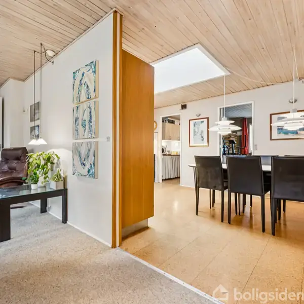 En stue og spisestue er adskilt af en halv væg, møbleret med en lænestol og et spisebord med seks stole; rummet er lyst med trægulv og loftpaneler.