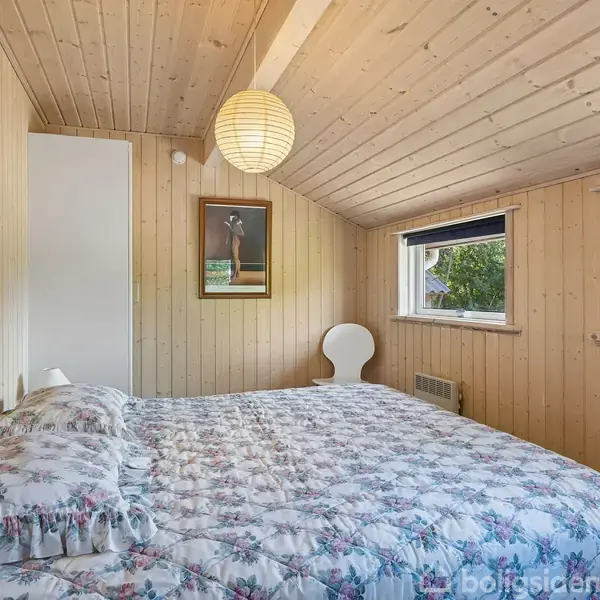 Soveværelse med blomstrende sengelinned på en dobbeltseng, træbeklædte vægge og loft, to små vinduer med udsigt til have, og billeder på væggene; loftslampe hænger centralt.