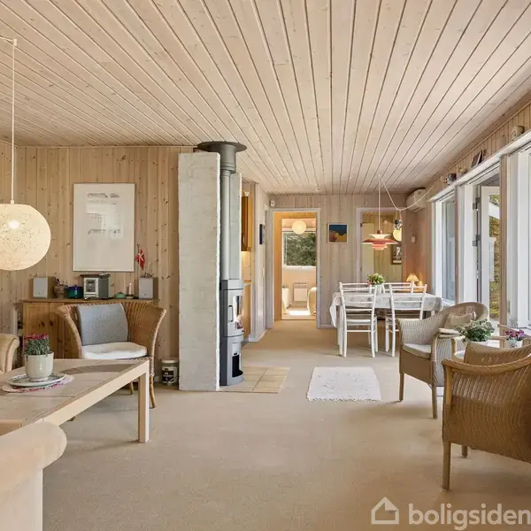 Stue med trævægge og loft, beige møbler, spisebord med stole i baggrunden, store vinduer til højre giver udsigt til en have. Pejs i midten af rummet.