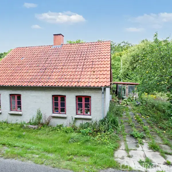 Et hvidt hus med rødt tegltag og røde vinduer står stille i en frodig have med grønt græs og buske. En gangsti fører til en tilbygning bagved.
