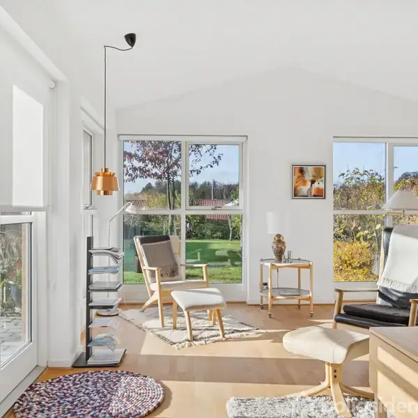 Et lyst stueområde med lænestole og små borde, indrammet af store vinduer, der viser en have udenfor. Lyse vægge og forskellige tæpper giver en hyggelig atmosfære.