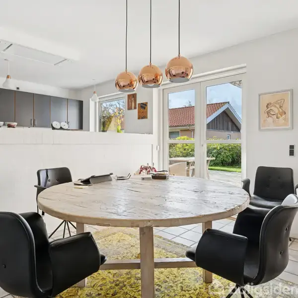 Rundt træbord med fire sorte stole står i et lyst køkken-alrum. Pendellamper hænger over bordet. Store glasdøre fører ud til en have og slipper dagslys ind.