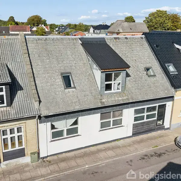 Rækkehus i en boligblok med skråt tag og ovenlysvinduer. Parkeret bil langs fortovet. Roligt byområde med omkringliggende træer og blå himmel i baggrunden.