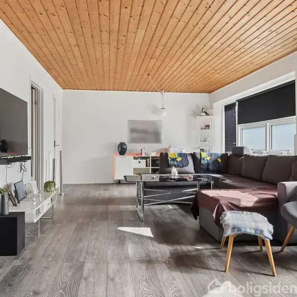 Stue med hjørnesofa og sofabord i glas, fladskærms-TV på væggen, hvidmalede vægge, træloft og gråt trægulv. Store vinduer med sorte gardiner tillader naturligt lys.