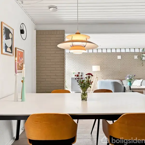 Spisebord med seks brune stole står i et lyst rum. Over bordet hænger en moderne lampe. Væggen er dekoreret med flere kunstværker. Til højre ses en stue med sofa.