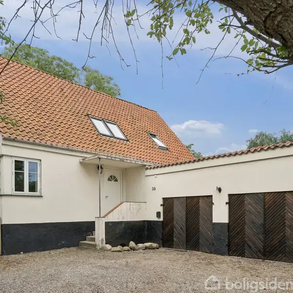 Et hvidt hus med rødt tegltag står stille i en haveomgivelser. To træbeklædte garageporte ved siden af hoveddøren. Adresse viser nummer 35. Træramme ses i forgrunden.