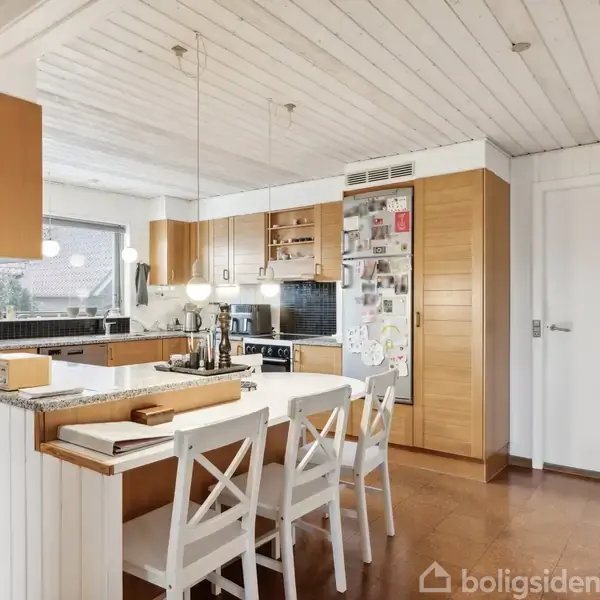 Et moderne køkken med træmøbler og en hvid spisebord med fire stole. Køleskabet er dekoreret med magneter og fotografier. Rummet har hængende lamper og et bredt vindue med udsigt.