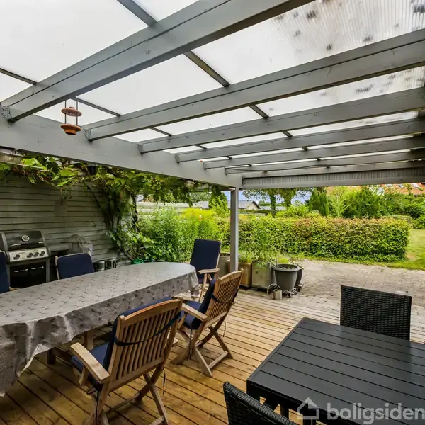 En overdækket terrasse med træmøbler om et bord med dug. En grill står i baggrunden ved hegnet. Terrassen åbner ud til en have med grønne buske og græs.