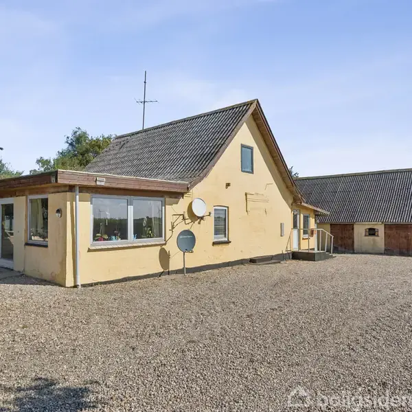 Et gulmalet hus med skråt tag står på en grusbelagt gårdsplads. Flere vinduer ses på bygningen, og der er satellitantenner monteret. Omgivet af træer og åbne marker.