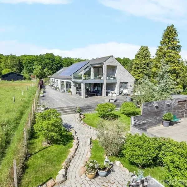 Et moderne hus med solpaneler ligger i en stor have med flisebelagt terrasse. En stensti fører gennem græsplænen, omgivet af træer og buske, beliggende i et landligt område.
