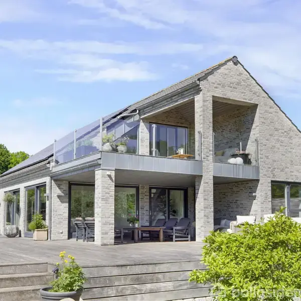 Et moderne hus i to etager med glasrækværk på altanen. Huset er omgivet af en træterrasse og grønne planter under en klar blå himmel.