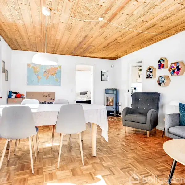 En stue med trægulv og træloft, viser et spisebord med hvide stole, en grå lænestol og sofa. På væggen hænger et verdenskort og hylder. En dør åbner til et andet rum.