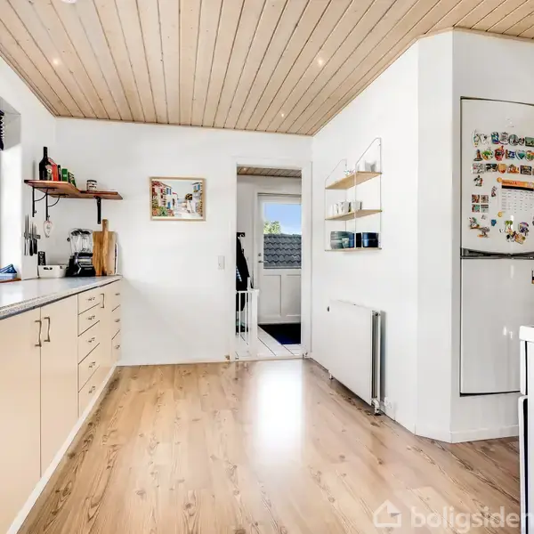 Køkken med lyse træskabe og granitbordplade indeholder køkkenmaskiner og planter ved vinduet. I midten fører en åben dør til en gang, mens køleskab med magneter ses til højre.