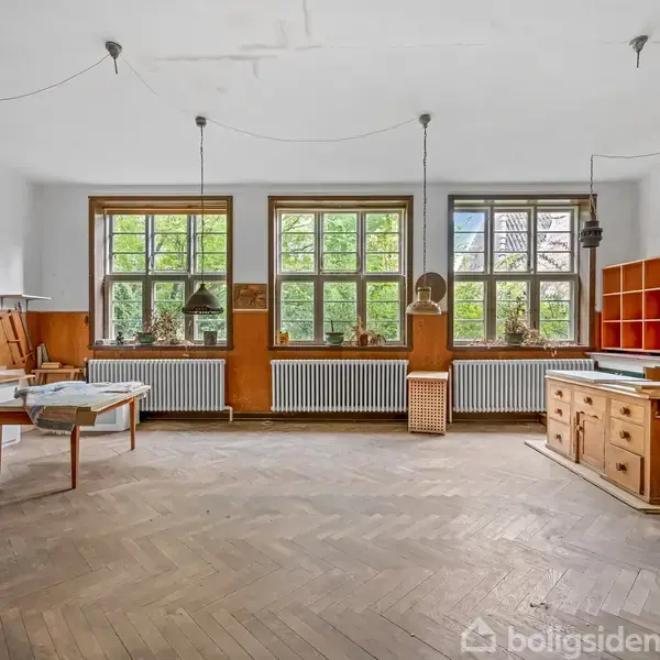 Et værksted er fyldt med træbord, hylder, vinduer og lamper. Store vinduer giver udsigt til grønne omgivelser udenfor. Gulvet er lagt med parket, og rummet har en afslappet atmosfære.