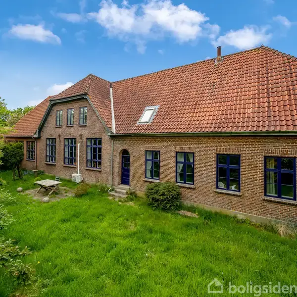 Stort murstenshus med rødt tegltag, omkranset af frodig have med træer og græs. Der står et havebord foran huset, som har blåmalede vinduer og døre.