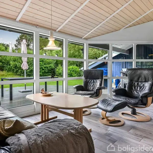 Stue med to sorte lænestole og et sofabord set fra en beige sofa. Store vinduer giver udsigt til en grøn have. Et fjernsyn er monteret på væggen.