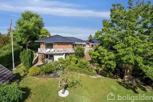 Et rødt murstenshus med sort tag står midt i en frodig have. Huset har en stor, hvid balkong, omgivet af træer og grønne planter under en blå himmel.