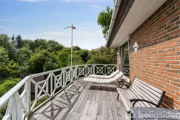 En træterrasse med to liggestole og en bænk vender ud mod en frodig have. Terrassen er ved siden af en murstensvæg under en klar, blå himmel.