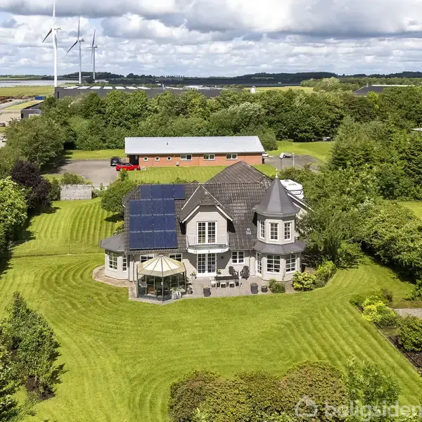 Et stort hus med solpaneler på taget ligger omgivet af en velplejet græsplæne og tæt beplantning. I baggrunden ses flere bygninger og vindmøller på en overskyet dag.