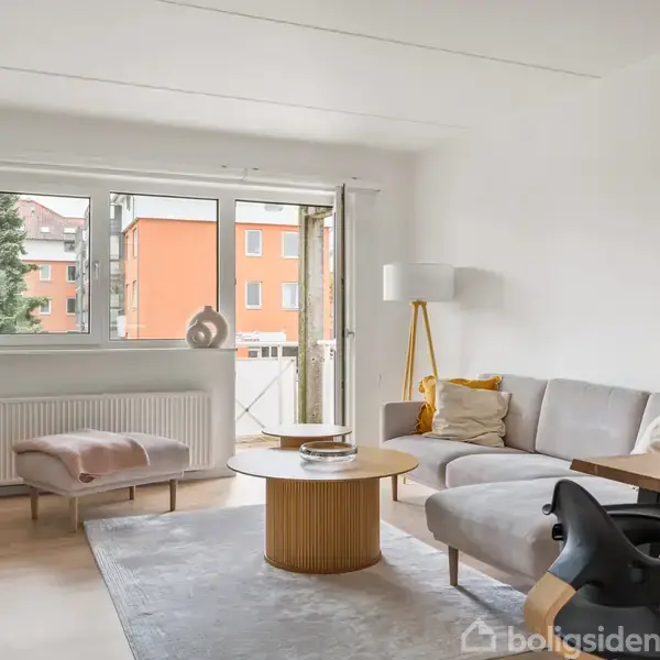 En lys stue med en grå hjørnesofa ved siden af et rundt træbord. En moderne gulvlampe står ved sofaen. Store vinduer giver udsigt til en rød bygning og et træ.