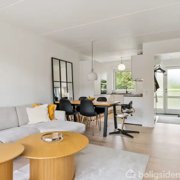 Lys stue med moderne gråt sofaarrangement og runde træborde. Spisebord med sorte stole ved siden af et åbent køkken. Store vinduer giver udsigt til en grøn have.