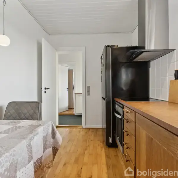 Køkkenskab i træ står til højre med køkkenbord og kogeplade. En grå stol står ved et bord dækket af en ternet dug. Bagved ses en åben dør i en lys gang.