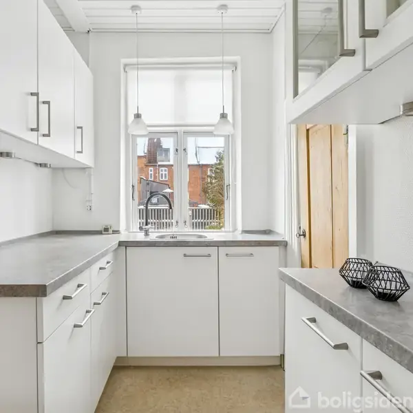 Køkken med hvide skabe og grå bordplader; to hængende lamper over vindue med udsigt til en bygning udenfor. Der er dekorative kurve på bordet.