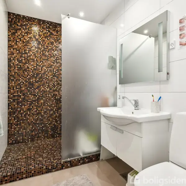 Badeværelse med toilet og vask. Brusekabinen har mosaikfliser i brune nuancer. Håndklæde hænger på døren. Vasken har spejl og tandbørster. Lyse fliser dækker resten af væggene.