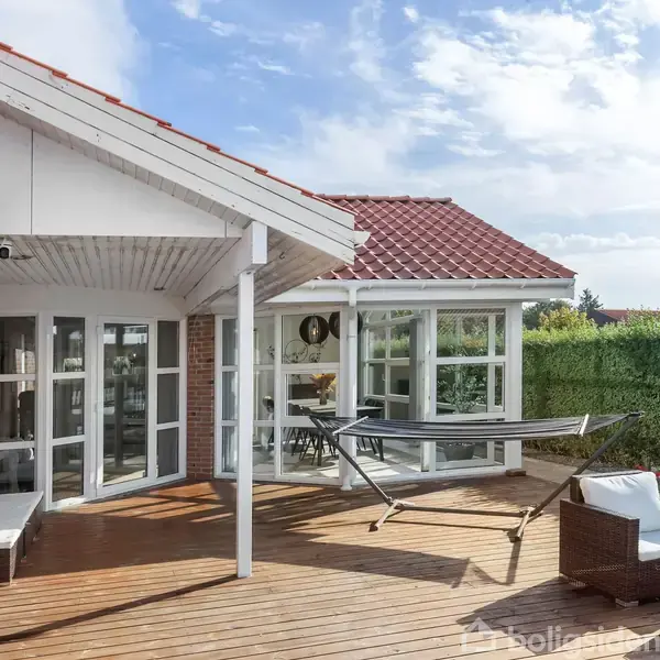 Terrasse i træ med hængekøje og havemøbler. Til venstre ses en grill, mens store vinduespartier på huset åbner ud til haven, omkranset af grønne hække under en delvis overskyet himmel.