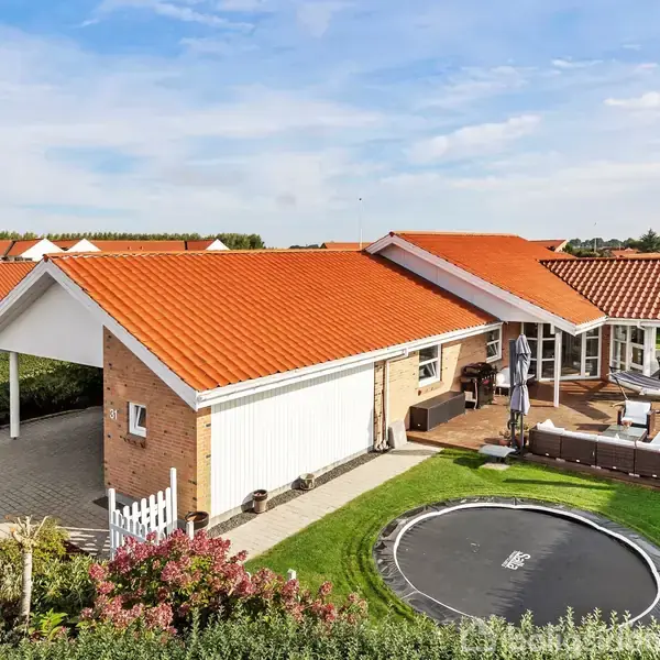 En moderne villa med rødt tegltag og garage til venstre; i baghaven er en terrasse med havemøbler og en trampolin omgivet af grønne hække og en klar blå himmel over.