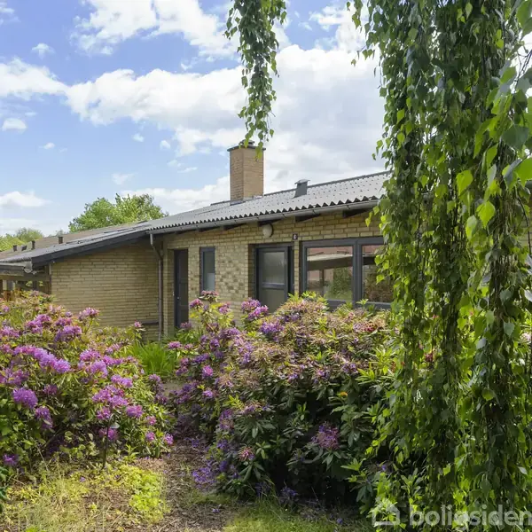 En murstensbygning med fladt tag står blandt farverige blomster og grønne hængeplanter i en velplejet have, under en blå himmel med spredte skyer.