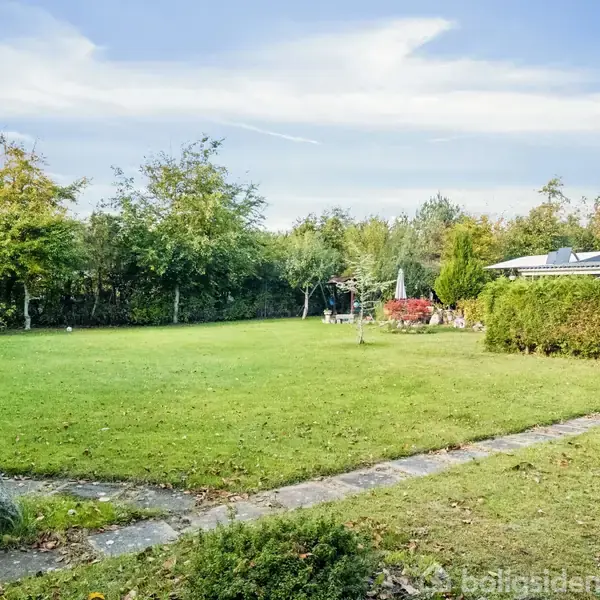 En stor, velplejet græsplæne med en flisebelagt sti ligger i en have omkranset af træer og buske. En terrasse med parasol og havemøbler er synlig til højre.