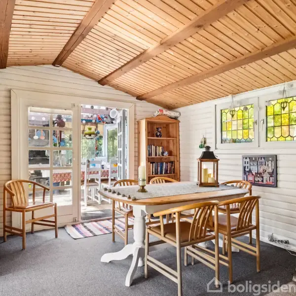 Spisebord med stole står i et lyst rum med træloft. Glasdøre fører til en terrasse. Væggen er dekoreret med farvede vinduer, en reol og en lampe.